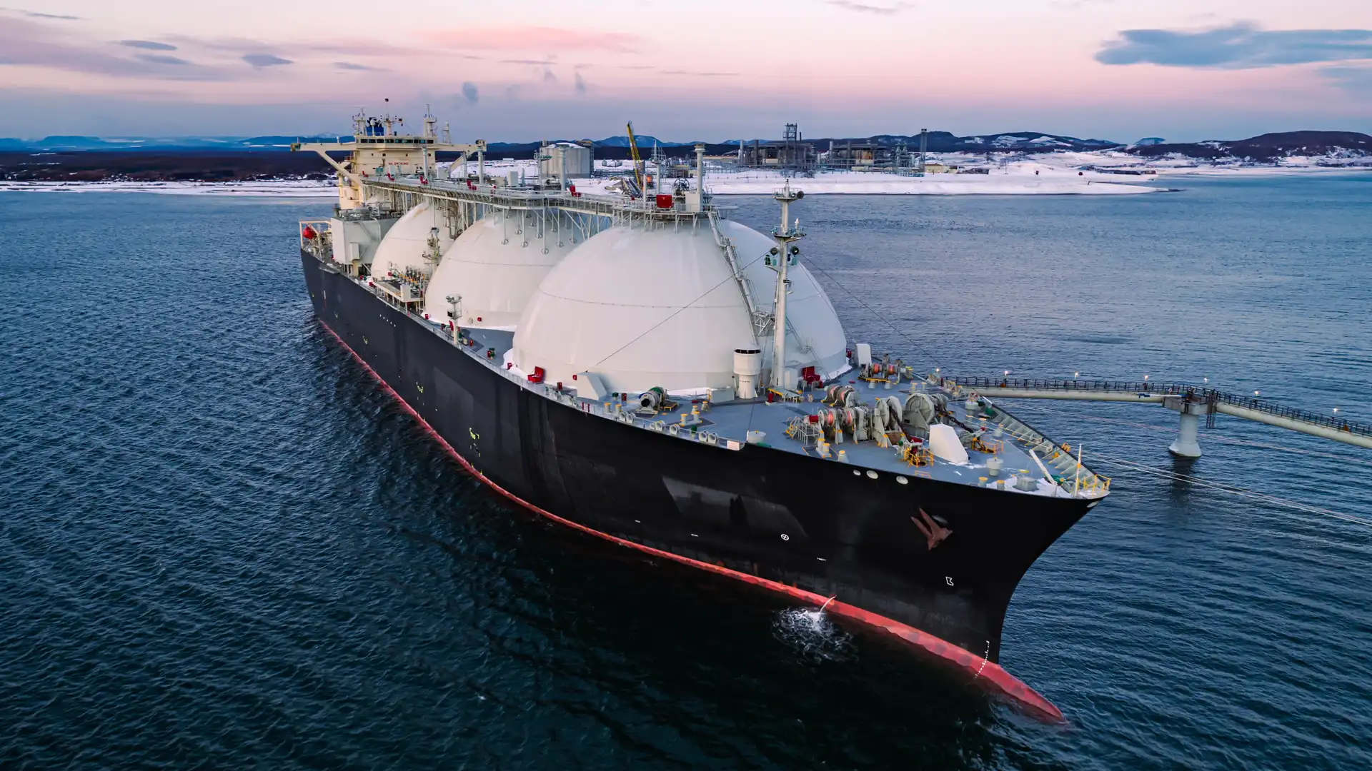 Large LNG tanker vessel at sea near a terminal, with spherical storage tanks visible on deck during sunset.